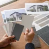 木製テーブルの上で、人物がグレー系の色見本を手に取り、住宅の外観写真や素材サンプルと見比べている