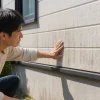 屋外でしゃがんだ男性が、縦筋状の汚れが付いた建物の外壁に手を触れて確認している