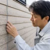 作業着姿の男性が、塗装の剥がれた住宅の外壁に手を当てて継ぎ目を確認している