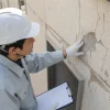 白いヘルメットと手袋を着けた作業員が、ひび割れて一部が剥がれた建物の外壁を手で確認している