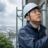 雨の中、白いヘルメットと紺色の上着を着た男性が足場から建物の外壁に手を触れて見上げている