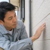 灰色の作業着を着た男性が屋外で建物の外壁に手を当て、表面の状態を確認している