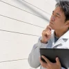 白い作業服の男性がタブレットを抱え、顎に手を当てながら建物の白い外壁を見つめている